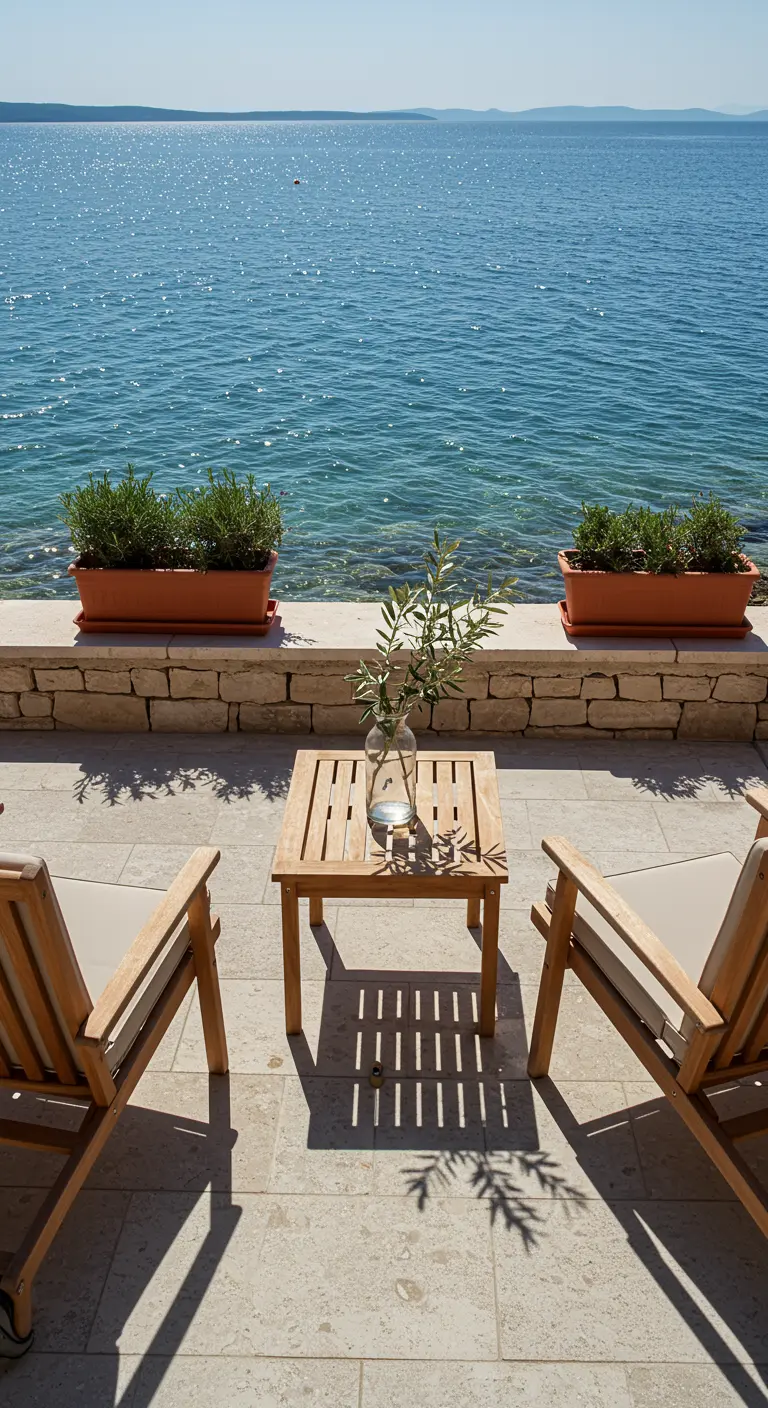 Two wooden chairs and a table facing the sea, with olive branches in a vase.